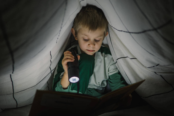 Little boy reading a book at night with lantern covered with blanket - Фото, зображення