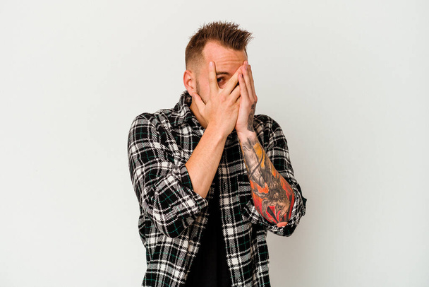 Young tattooed caucasian man isolated on white background blink through fingers frightened and nervous. - Fotó, kép