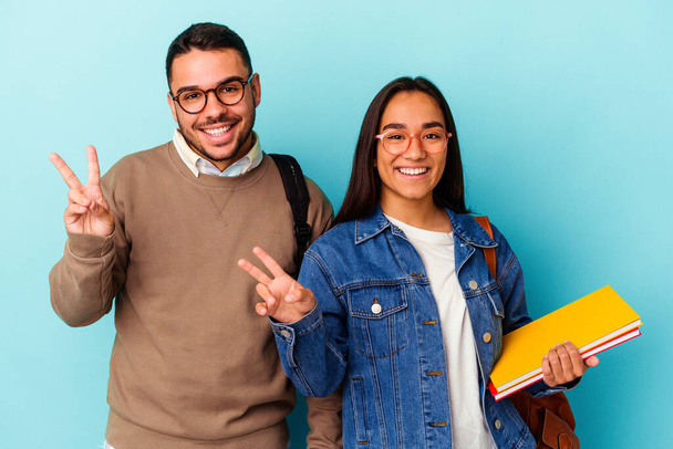 Young mixed race student couple isolated on blue background joyful and carefree showing a peace symbol with fingers. - Foto, imagen