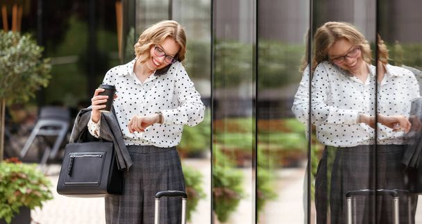 Улыбающаяся деловая женщина спешит в аэропорт, стоя на городской улице с чемоданом и унося с собой кофе, глядя на часы под рукой при вызове такси по мобильному телефону. Концепция деловой поездки - Фото, изображение