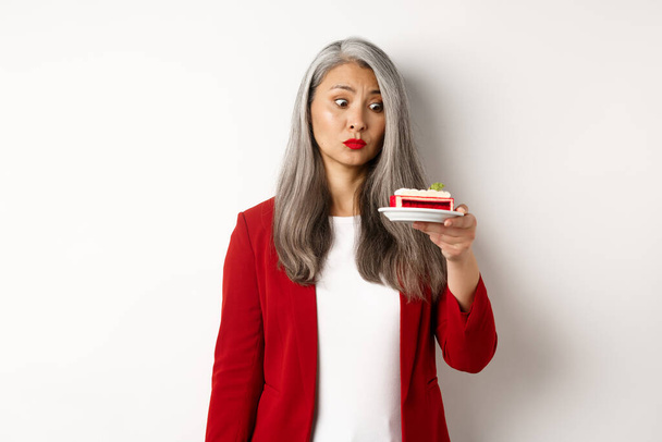 Elegant senior woman in red blazer wants take bite of sweet cake, looking with tempted face at dessert, standing over white background - Photo, Image