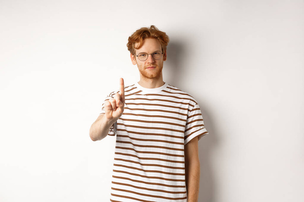 Junger Mann mit roten Haaren und Brille, der den Finger in Prohibition, Tabu-Geste, Missbilligung und Stopp etwas schüttelt, vor weißem Hintergrund stehend - Foto, Bild