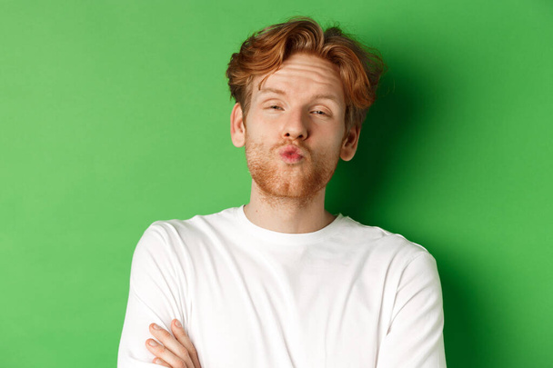 Emotions and fashion concept. Silly redhead guy with beard pucker lips and leaning for kiss, standing over green background - Foto, imagen