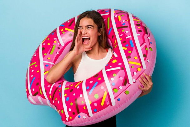 Young caucasian woman with inflatable donut isolated on blue background shouting and holding palm near opened mouth. - 写真・画像