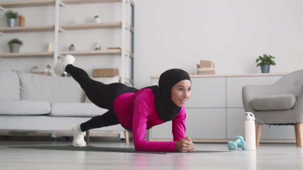 Posilovací cvičení. Mladá středovýchodní islámská dáma nosí hidžáb stojící v poloze prken a zvedá nohy - Záběry, video
