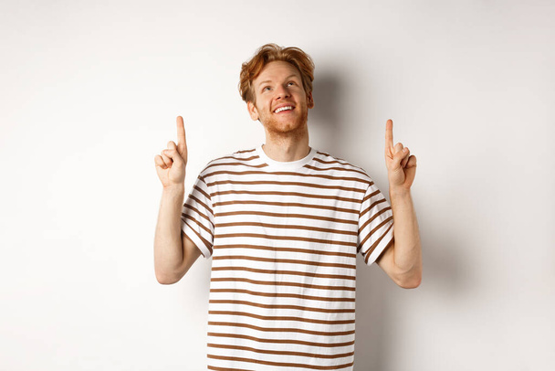 Happy young man with red hair and beard pointing, looking up with dreamy smile, checking out special promo offer, white background - Photo, Image