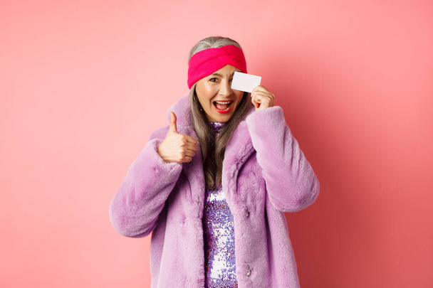 Zakupy i koncepcja mody. Piękna azjatycka kobieta w średnim wieku pokazuje plastikową kartę kredytową na twarzy i pokazuje kciuki do góry z podekscytowany, szczęśliwy wyraz twarzy, różowe tło - Zdjęcie, obraz