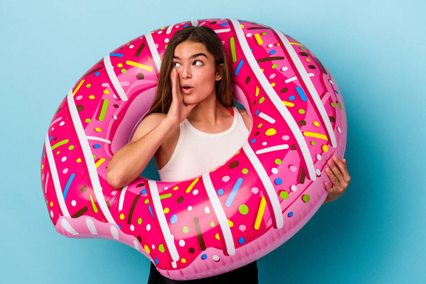 Young caucasian woman with inflatable donut isolated on blue background is saying a secret hot braking news and looking aside - Photo, Image