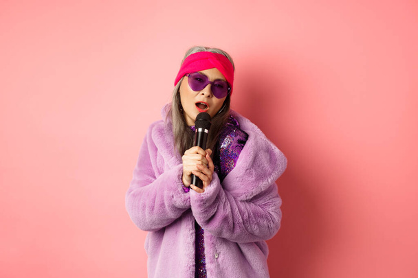 Fashion concept. Stylish senior asian woman singing karaoke, perform on stage with microphone, wearing trendy sunglasses and purple faux fur coat, pink background - Foto, afbeelding