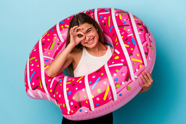 Young caucasian woman with inflatable donut isolated on blue background excited keeping ok gesture on eye. - Photo, Image