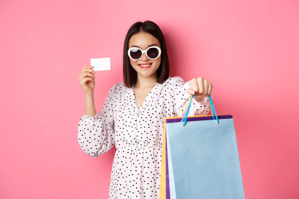 Güneş gözlüklü güzel Asyalı kadın alışverişe çıkıyor, çantalarını tutuyor ve kredi kartını gösteriyor, pembe arka planda duruyor. - Fotoğraf, Görsel