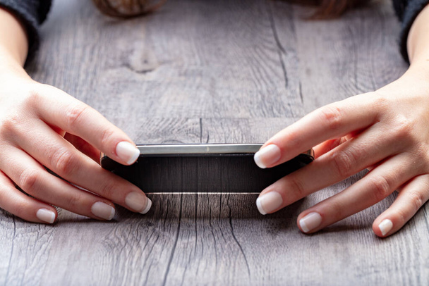 smartphone horizontaal ondersteund door twee vrouwelijke handen op een grote houten plank, de gemanicuurde handen hebben de Franse manicure - Foto, afbeelding