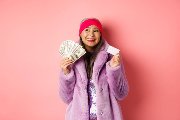Zakupy i koncepcja mody. Modna azjatycka seniorka marząca o sklepach i sprzedaży, trzymająca pieniądze w dolarach i plastikową kartę kredytową, różowe tło - Zdjęcie, obraz