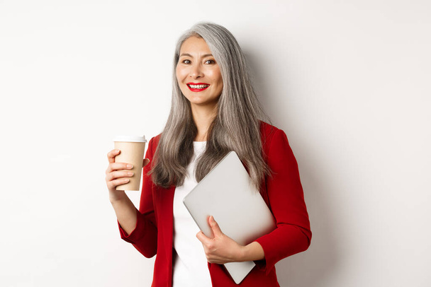 Business. Successful asian businesswoman with grey hair, wearing red blazer, drinking coffee and standing with laptop in hand, white background - Photo, Image