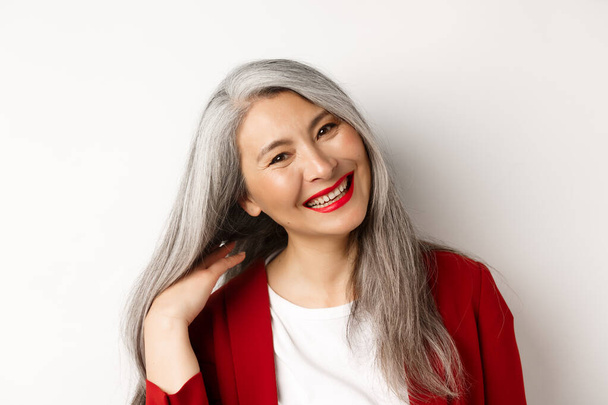 Concepto de belleza y envejecimiento. Acercamiento de mujer mayor asiática con labios rojos, pelo largo y saludable gris, sonriendo a la cámara, de pie sobre fondo blanco - Foto, Imagen