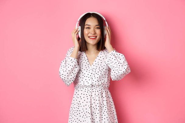 Romantická asijská žena se šťastně usmívá, poslouchá hudbu ve sluchátkách a dívá se do kamery, stojí nad růžovým pozadím - Fotografie, Obrázek
