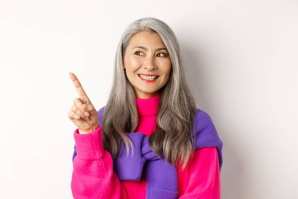 Gros plan de belle asiatique senior femme souriant, regarder et pointant publicité coin supérieur gauche, vérifier promo offre, debout sur fond blanc - Photo, image