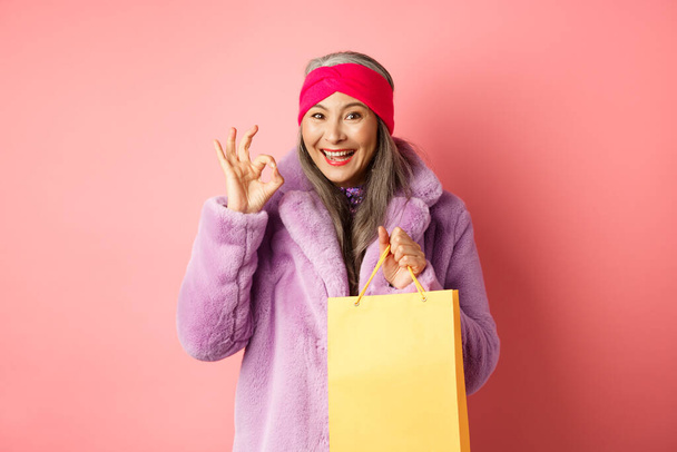 Compras y moda. Mujer mayor asiática feliz en ropa elegante sosteniendo bolsa de papel de la tienda, mostrando signo OK, recomendando tienda, fondo rosa - Foto, Imagen
