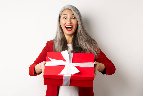 Sorprendido y agradecido abuela asiática recibiendo regalo en caja roja, mirando sorprendido y agradecido, de pie sobre fondo blanco - Foto, imagen
