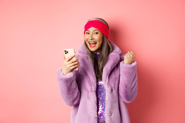 Achats en ligne et concept de mode. Heureux asiatique senior femme gagnant prix dans Internet, tenant téléphone mobile et faisant poing pompe, cri de joie, debout sur fond rose - Photo, image