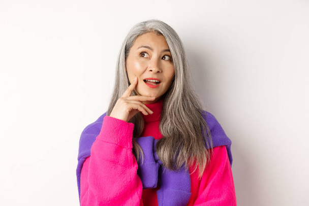 Primo piano di bella donna anziana asiatica in abiti eleganti, fare piani, guardando da parte e pensando, in piedi su sfondo bianco - Foto, immagini