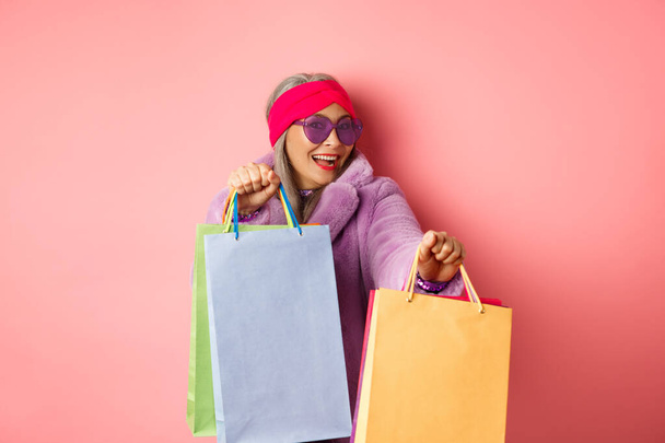 Пожилая женщина в модной одежде танцует во время шопинга на распродажах, держа в руках магазинные бумажные пакеты и развлекаясь на розовом фоне - Фото, изображение