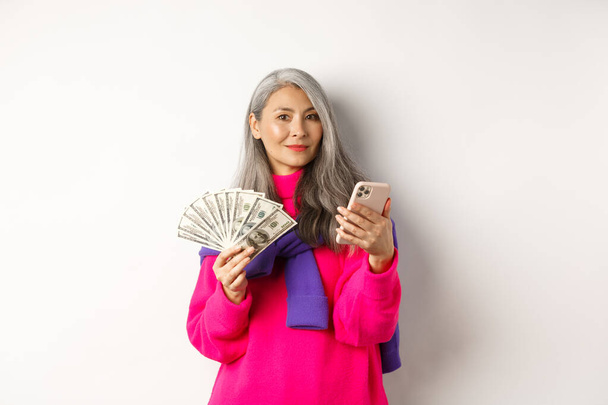 Moderna signora anziana asiatica in possesso di denaro e smartphone, ordinare qualcosa online, in piedi su sfondo bianco - Foto, immagini