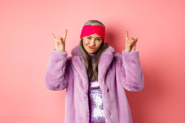 Cool mujer madura asiática en abrigo de piel sintética de moda, mostrando señales de cuernos de rock-n-roll y sonriente descarada en la cámara, divirtiéndose, de pie sobre fondo rosa - Foto, Imagen