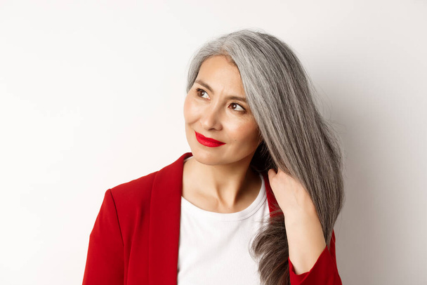 Conceito de beleza e cuidados com o cabelo. Close up de elegante asiático sênior mulher mostrando brilhante e saudável cabelos grisalhos, sorrindo e olhando para o lado, fundo branco - Foto, Imagem