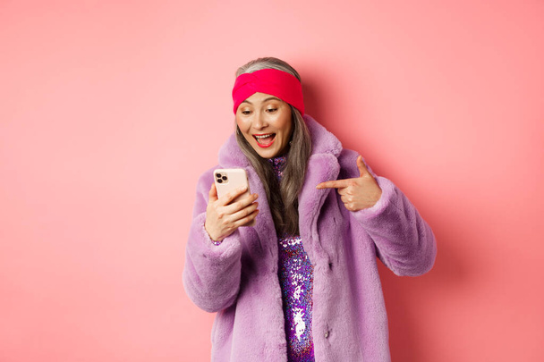 Online winkelen en mode concept. Cool aziatische senior vrouw controleren promotie op mobiele telefoon, wijzende vinger naar smartphone scherm en glimlachen blij, roze achtergrond - Foto, afbeelding