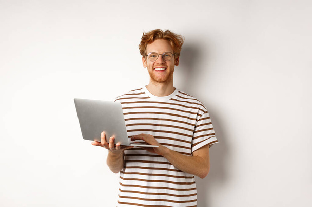 Sonriente joven que trabaja en el ordenador portátil y se ve alegre, de pie sobre fondo blanco - Foto, Imagen