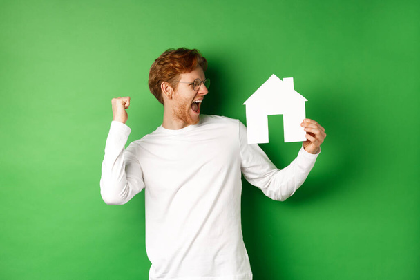 Nemovitosti. Veselý zrzek muž v brýlích slaví, kupuje dům, ukazuje papír doma střih a křičí radostí, stojící nad zeleným pozadím - Fotografie, Obrázek