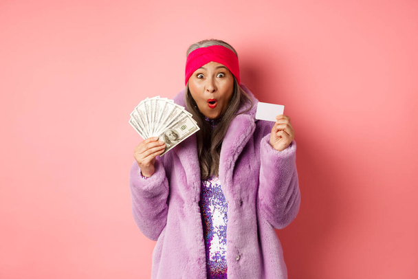 Zakupy i koncepcja mody. Szczęśliwy starszy Azji kobieta posiadające pieniądze w dolarach i karty plastikowej, patrząc zdumiony i ekstatyczny, różowy tle - Zdjęcie, obraz