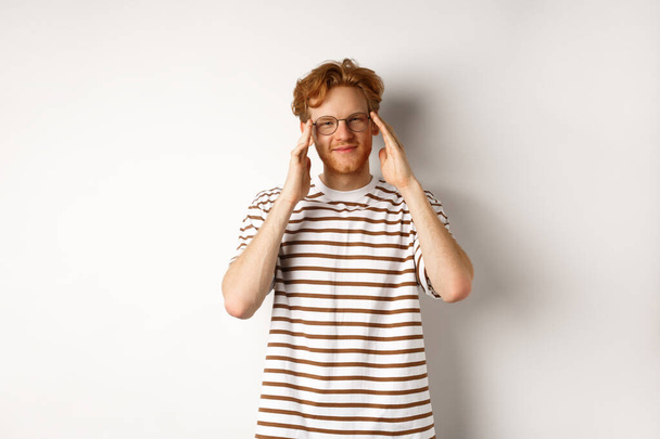 Jóképű fiatalember hipszter vörös frizurával és szemüveggel, mosolyog a kamera előtt, fehér háttér felett áll - Fotó, kép