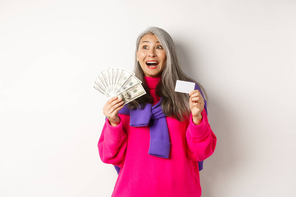 Concept de shopping. Vieille femme asiatique heureuse avec les cheveux gris, regardant fasciné au coin supérieur gauche, montrant les dollars de carte de crédit et d'argent en plastique, fond blanc - Photo, image