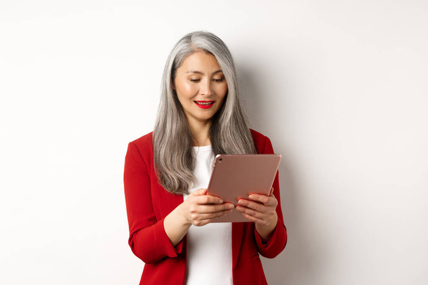 Negócios. Empresária sênior bem sucedida trabalhando com tablet digital, tela de leitura e sorrindo, de pé em elegante blazer vermelho sobre fundo branco - Foto, Imagem