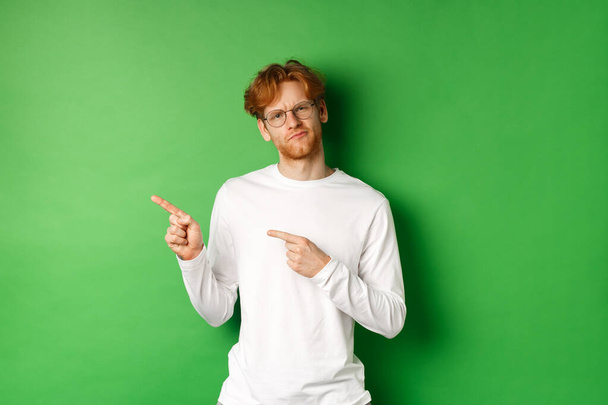 Zweifelhafter Rotschopf mit Brille zeigt mit dem Finger nach links, blickt zögerlich und unsicher in die Kamera, steht über grünem Hintergrund - Foto, Bild