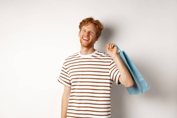 Cheerful bearded man with red hair enjoying shopping, holding bag over shoulder and smiling, standing over white background - Φωτογραφία, εικόνα