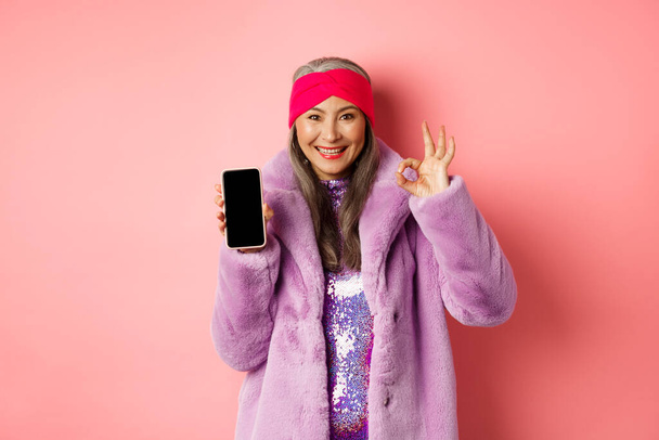 Online winkelen en mode concept. Modieuze senior aziatische vrouw tonen blanco zwarte smartphone scherm en OK teken, demonstreren promotie, roze achtergrond - Foto, afbeelding
