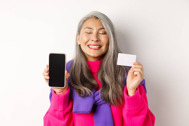 Achats en ligne. Heureuse et ravie asiatique femme âgée souriant de joie, montrant écran smartphone vierge et carte plastique, acheter sur Internet, fond blanc - Photo, image
