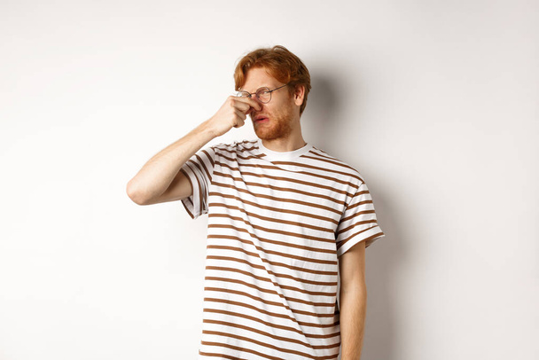 Young man with red hair staring at camera and shut nose from bad smell, looking disgusted as something reek, white background - Photo, Image