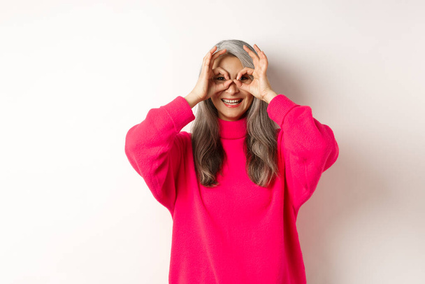 Feliz y divertida asiática senior mujer haciendo binoculares de mano, mirando a través de los dedos y sonriendo, de pie en suéter rosa sobre fondo blanco - Foto, Imagen