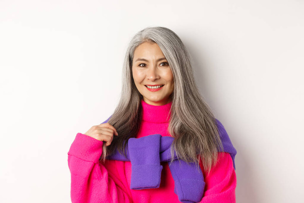 Close-up of beautiful asian senior woman in trendy pink sweater, smiling cheerful at camera, standing over white background - Zdjęcie, obraz