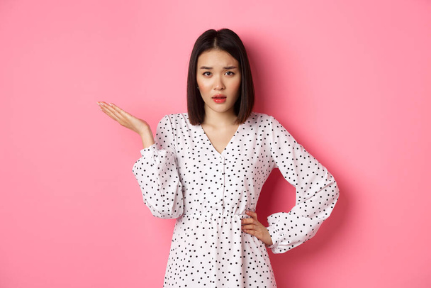 Careless asian woman raising hand and shrugging, staring at camera unbothered and questioned, so what gesture, standing over pink background - Photo, Image