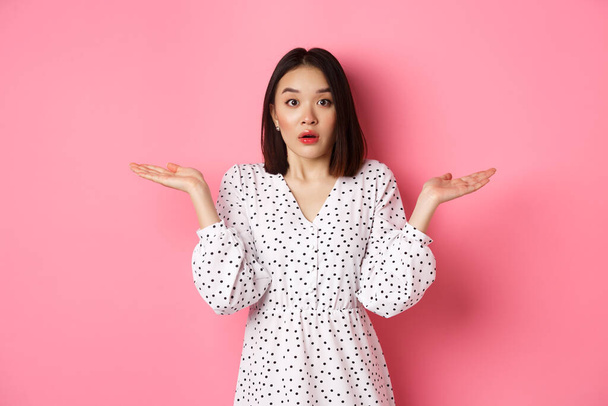Confused and clueless asian girl shrugging, staring at camera puzzled, dont know what to do, spread hands sideways, standing over pink background - Photo, Image