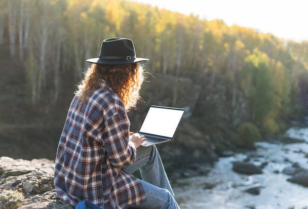 Young beautiful woman with curly hair in felt hat and plaid shirt using laptop on view background of mountains and river, hiking on autumn nature, concept of freelance - Foto, Bild