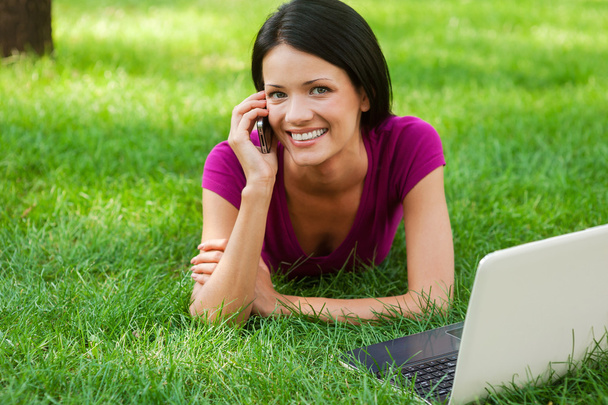 Woman talking on phone lying in grass with laptop - Foto, Imagen