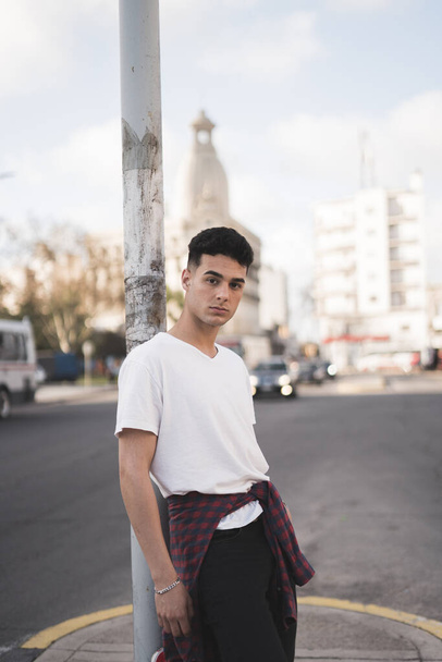 A vertical shot of a young stylish male in a white t-shirt leaning against a street lamp post - Фото, изображение