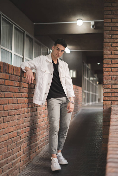 A vertical shot of a young stylish caucasian man in a white jacket leaning on a brick wall - Valokuva, kuva