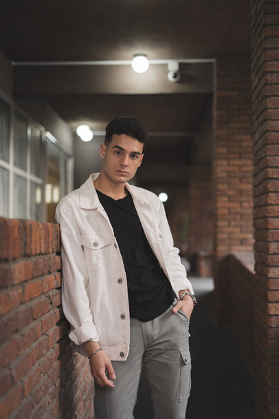 A vertical shot of a young stylish caucasian man in a white jacket leaning on a brick wall - Photo, image